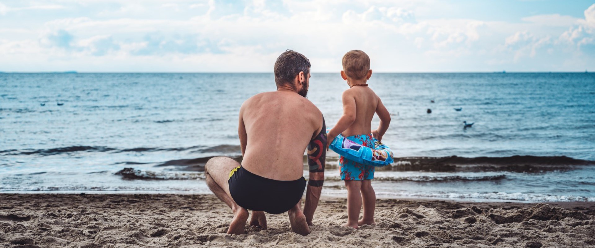 We see a father and son on the beach looking out at the sea in the image.